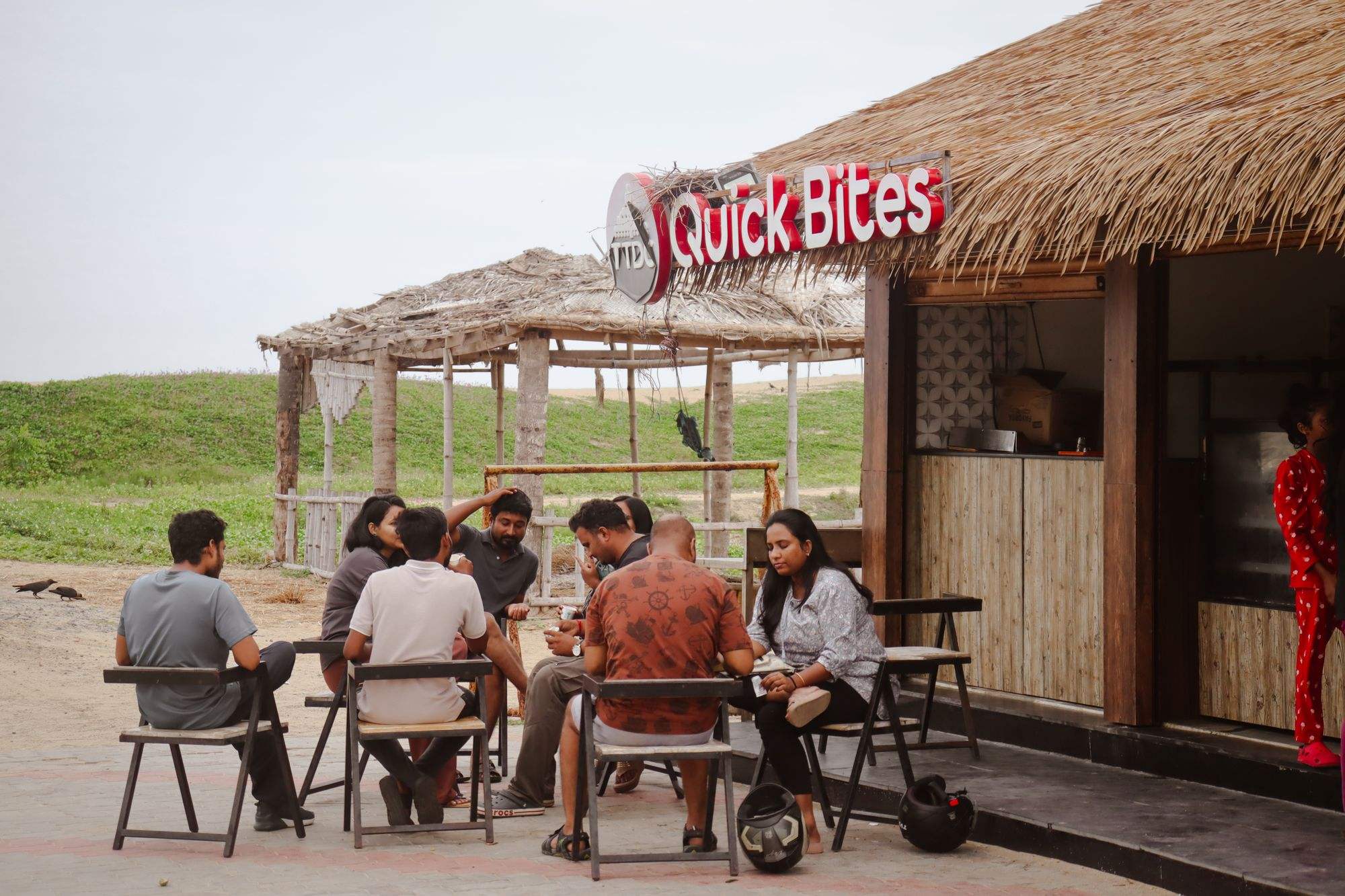 A group of friends sitting and talking at a beach snack shack, motorbike helmets on the ground beside them.