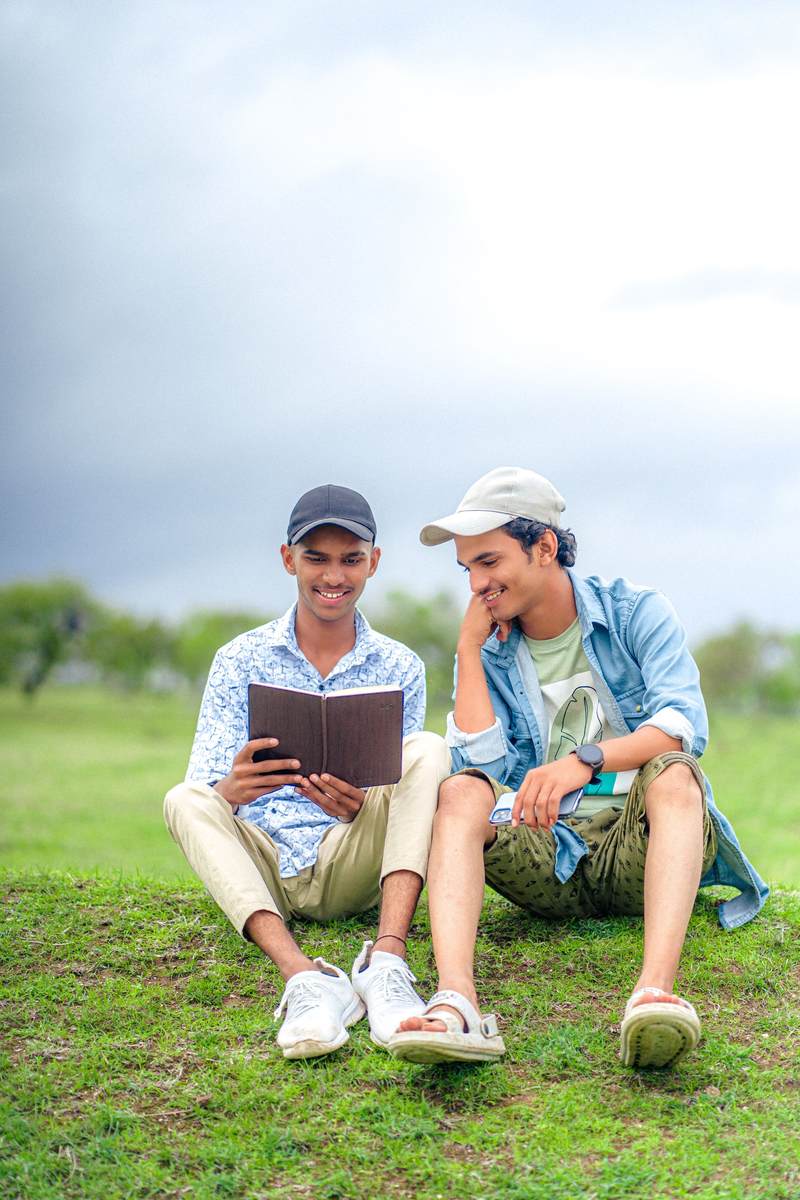 Two friends sitting on the grass in a park, one reading aloud from a book.